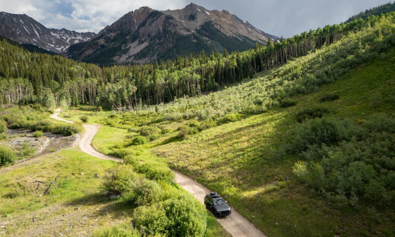 Gray Audi SUV overhead view driving on dirt road with green hills and distant mountains.