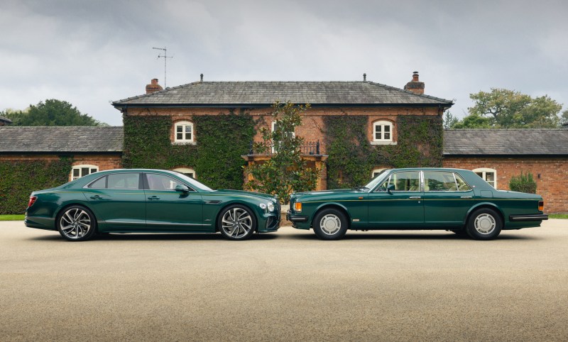 Bentley 2025 Flying Spur Speed and 1985 Turbo R facing each other on a driveway in front of brick buildings.