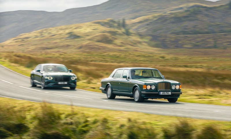 Bentley 2025 Flying Spur Speed and 1985 Turbo R at speed on a winding road in open country.