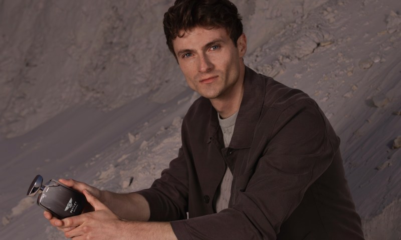 A person wearing a dark brown shirt holding a bottle of Bentley Become Zenith fragrance.